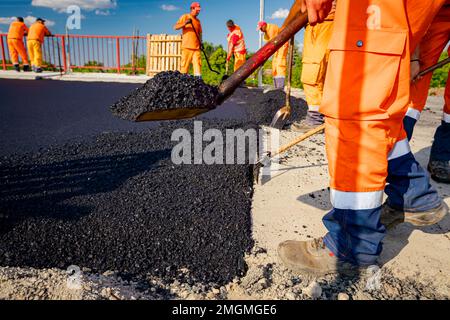 Few workers are using shovels to level, set up layer of fresh tarmac to right measures, pouring hot asphalt. Stock Photo