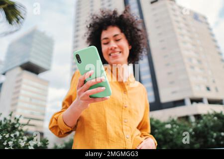Focus on smartphone. Young hispanic woman typing on cellphone and smiling outdoors. Unfocused happy person using a mobile phone. High quality photo Stock Photo