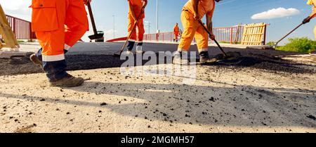 Few workers are using shovels to level, set up layer of fresh tarmac to right measures, pouring hot asphalt. Stock Photo