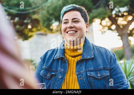 A happy and confident non-binary curvy woman sits in an outdoor bar. Smiling in this half-body portrait embracing her unique identity and celebrating Stock Photo