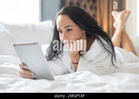 A good-looking brunette is lying on the bed in a bathrobe and using a tablet for a dating site. Stock Photo