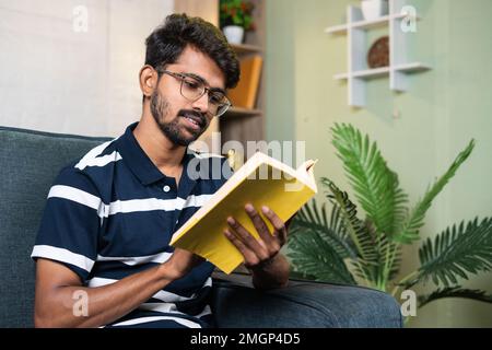 serious young indian student reading book during free time at home - concept of education, hobbies and hard worker. Stock Photo