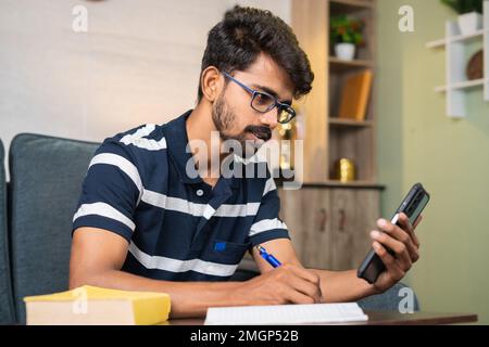 Young man using mobile phone for writing notes at home while listening online class - concept of e-learning, dedication and intelligence. Stock Photo