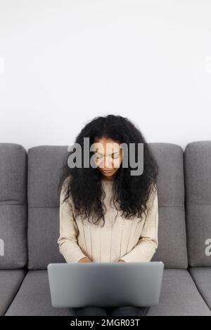 BIPOC woman works on laptop on couch at home. POC female person doing distant work online with modern notebook computer Stock Photo