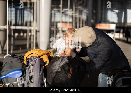 Fatherat comforting his crying infant baby boy child tired sitting on top of luggage cart in front of airport terminal station while traveling wih family Stock Photo