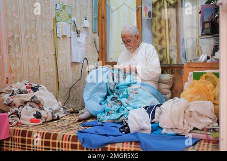 Fez, Morocco - old male tailor sews a bright blue abaya inside his shop in Fes el Bali. Seamster sits on a checkered surface with pillows and fabrics. Stock Photo