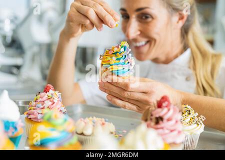 happy women in pastry bakery as confectioner sprinkles candies topping on a colorful muffin by hand Stock Photo