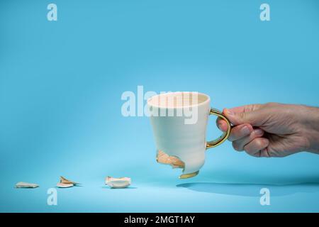 Man holding broken tea cup isolated on blue background. Close up of arm holding cracked coffee mug Stock Photo