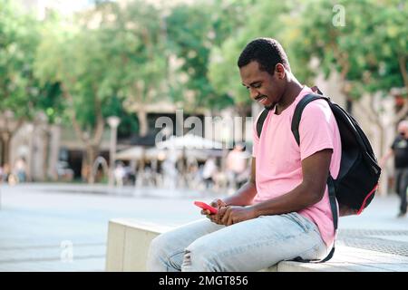 Man with backpack using a mobile phone while sitting on a bench outdoors. Stock Photo