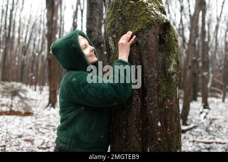 Happy woman in winter forest. Female portrait in snow park. Smiling woman in eco fur jacket touch with hand tree trunk. Stock Photo