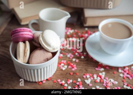 Macaroons in a bowl, a cup of coffee, a milk jug on a background of small hearts on a wooden background Stock Photo