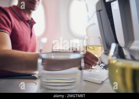 Man enjoying drink during flight. Passenger holding glass of white wine against airplane window. Stock Photo