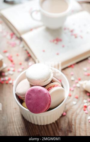 Macaroons in a bowl and a cup of coffee, a book on the background of small hearts on a wooden background, top view Stock Photo