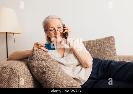 Aged woman making a call and holding a credit card. Senior female making an online order. Stock Photo