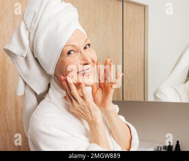 Smiling senior woman with a towel on her head massaging her face and looking at camera Stock Photo