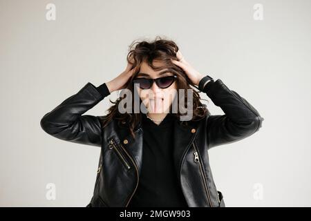 Brunette with curly hair, glasses and leather jacket shows her tongue isolated Stock Photo