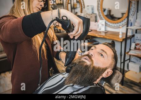 Female hairdresser using the hair dryer to straighten his long beard - Hipster man at barbershop salon getting beard and hair cut - Barber shop concep Stock Photo