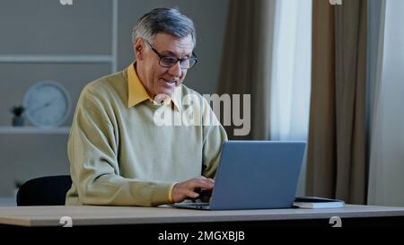 Old Caucasian man with computer work indoors typing chatting writing book online text. Elderly mature senior businessman worker entrepreneur Stock Photo