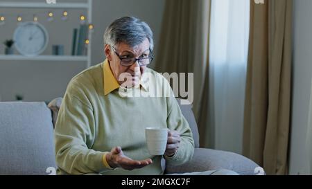Old elderly mature grandfather man sitting at home couch watching TV drinking hot tea from cup supporting team in online sports competition frustrated Stock Photo