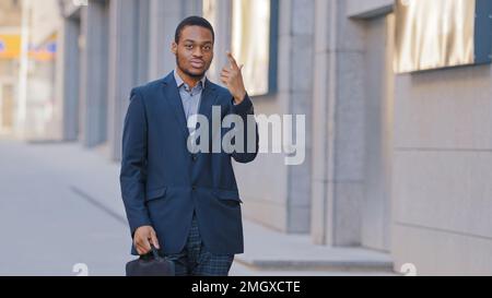 Confident African American serious man employee entrepreneur businessman ethnic male guy worker manager with suitcase in business suit show number one Stock Photo