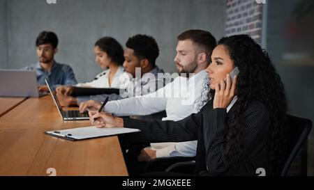 Multiracial employees sitting in conference room coworking work at table. Hispanic businesswoman CEO manager talking on mobile phone writing notes Stock Photo