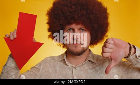 Upset man with afro hairstyle showing red arrow pointing down, concept of downgrade, unsuccessful business, fall of stock market money exchange rate, bankruptcy fail. Hipster guy on yellow background Stock Photo