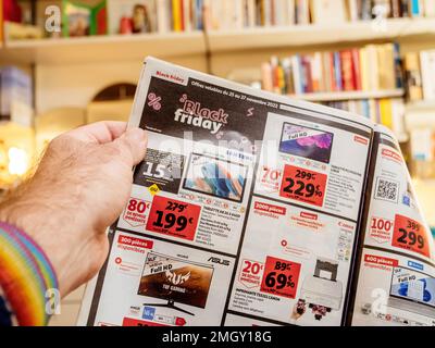 Paris, France - Dec 2, 2022: Man reading Auchan France leaflets supermarket chain selling furniture, electronics home object special black friday prices with Vitsoe 606 shelves in background Stock Photo