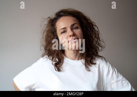 Portrait young caucasian curly woman wearing white t-shirt looking at camera against plain background. simplicity Stock Photo