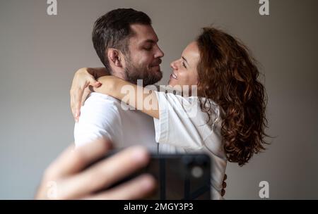Young adult couple wearing white tee shirts hugging taking selfie and smiling, couple in love. Stock Photo