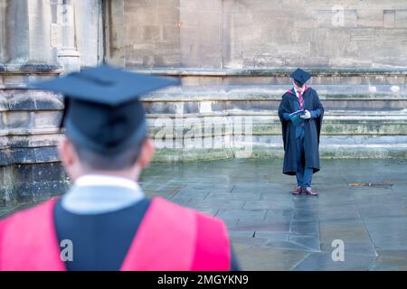 A young University graduate is shown texting on a mobile phone having just left a graduation ceremony. He's wearing a gown and  Mortar Board hat Stock Photo