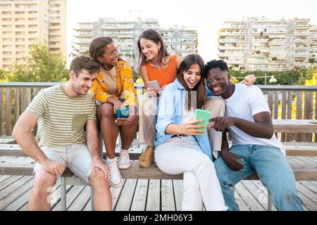 Group of young people standing together while sitting in wooden bench in the city - Funny multiethnic students watching their smartphones in the Stock Photo