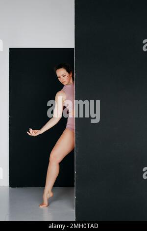 A pretty young woman peeks out from behind a black screen. Studio portrait of a young female gymnast in pink shorts and top. Graceful body movements. Stock Photo