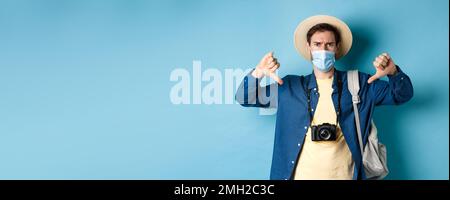 Covid-19 and summer holidays concept. Disappointed tourist in medicla mask frowning, showing thumbs down, negative feedback about travel, standing on Stock Photo