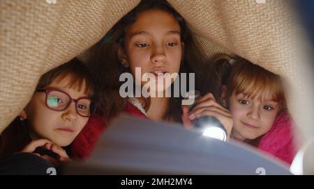 Teen girl and toddler child sisters under blanket plaid covers with flashlight reading interesting fairytale story book using flashlight. Female three siblings children kids friends in wigwam at home Stock Photo