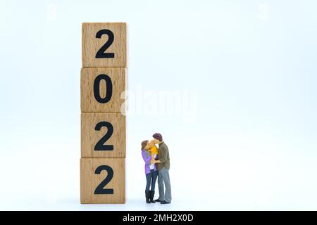 2022 New Year and Family Concept. Closeup of parent and child miniature figures hug and kissing with stack of wooden number block on white background. Stock Photo