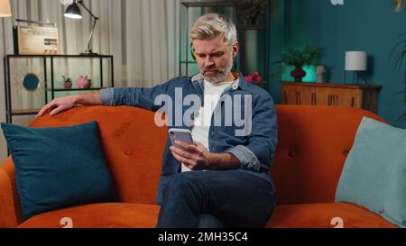 Bearded middle-aged man sitting on sofa uses mobile phone smiles at night home apartment. Mature guy texting share messages content on smartphone social media applications online, watching relax movie Stock Photo
