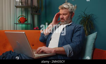 Tired middle-aged man freelancer use laptop, suffering from headache problem tension and migraine, stress at home. Mature guy works on notebook, sends messages, makes online purchases, watching movies Stock Photo