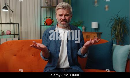 What. Why. Middle-aged man raising hands in indignant expression asking reason of failure demonstrating disbelief irritation by troubles at home apartment. Mature guy in evening room sitting on sofa Stock Photo