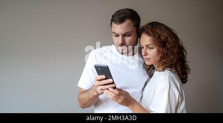 Young couple with blank expression wearing white t-shirt looks at screen of mobile phone against plain background Stock Photo
