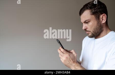 Serious adult bearded man in white t-shirt holding mobile phone in hands and looking attentively at screen of phone against plain background. Stock Photo