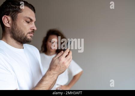 Bearded man reading messages in his mobile phone woman on background watching her husband. Stock Photo