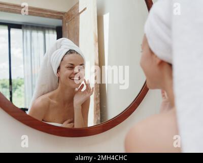 A beauty woman stands in front of a mirror after a shower in a towel on her head looks at her reflection and does a facial massage applies a day cream Stock Photo