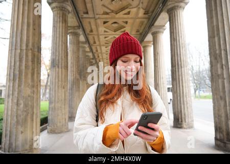 Mobile broadband and people. Smiling redhead 20s girl with backpack, uses smartphone on street, holds mobile phone and looks at application Stock Photo