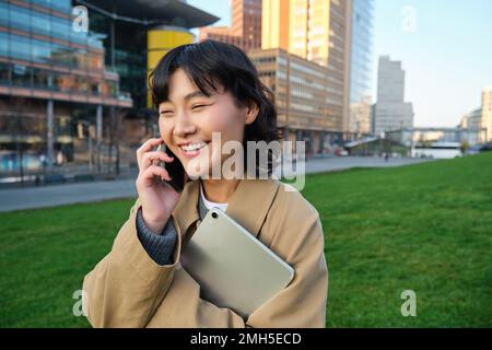 Stylish smiling girl, university student in trench coat, holds tablet, talks on mobile phone, has conversation over telephone and looks relaxed Stock Photo