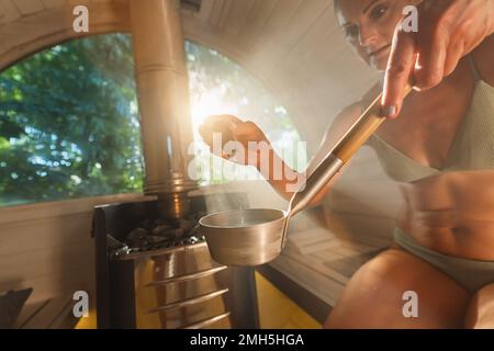 Woman dripping essential aromatic oil from a dropper bottle in to water for a trowel in a wooden barrel sauna in norway. Steam, spa and wellness Stock Photo