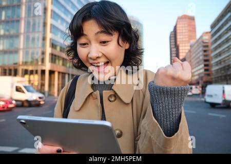 Enthusiastic asian girl with digital tablet, smiling and looking amazed, happy with something good, holding digital tablet, standing on street Stock Photo