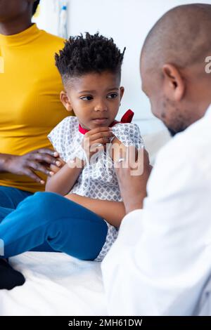 Vertical of african american male doctor vaccinating boy patient, with mother Stock Photo