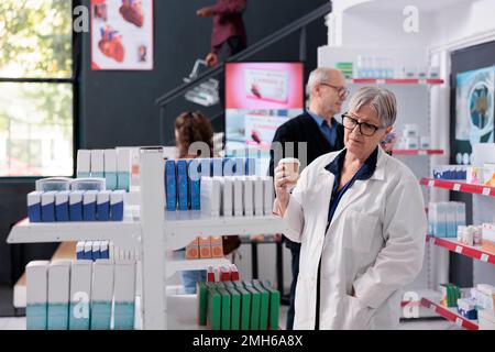Senior pharmacist holding cup of coffee while looking at drugstore shelves full with vitamins and pills, supplements. Diverse clients doing healthcare shopping session buying medication Stock Photo