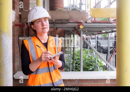 A concerned concentrated civil engineer woman surveying and supervising a construction site with scaffolding, wearing a hard hat and orange ppe Stock Photo
