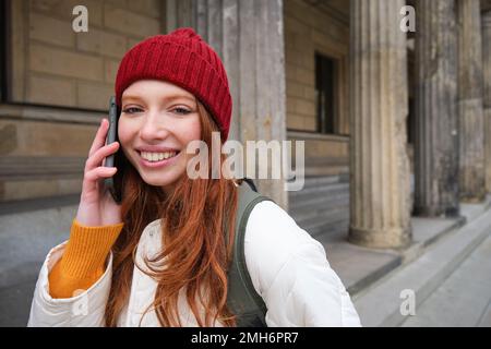 Smiling redhead female tourist talks on mobile phone and walks around city. Happy student in red hat calls friend, stands on street and uses Stock Photo